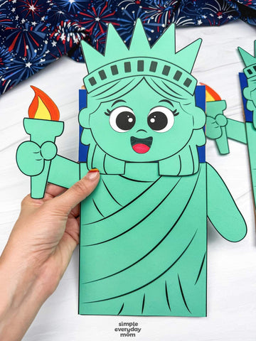 hand holding Statue of Liberty paper bag puppet craft on white background with fireworks fabric on top