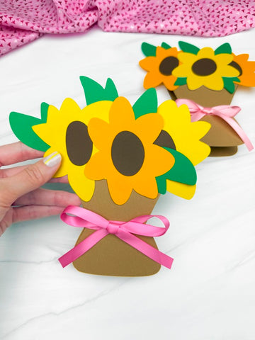 hand holding sunflower card craft with 2nd one in the background. White background with pink fabric on top. 