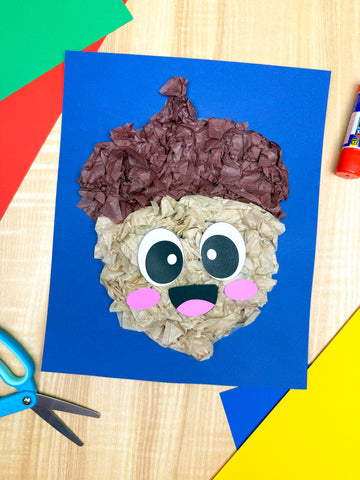 Children's acorn project with a face made from crumpled paper on blue paper, surrounded by colorful shapes and a glue stick.