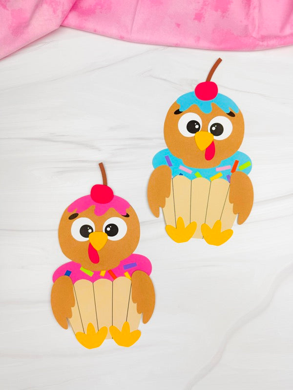 Two colorful turkeys disguised as cupcakes on a white surface with a pink fabric background.