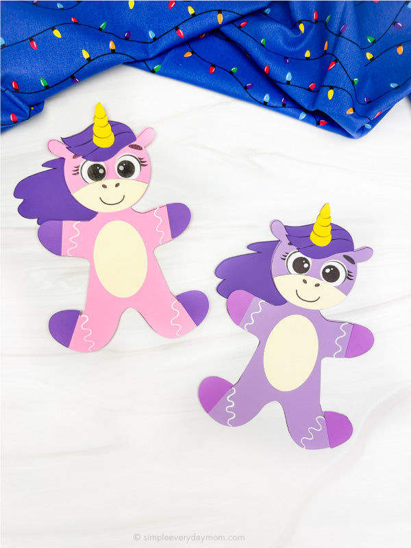 Two colorful paper unicorn gingerbread disguise crafts on a white background with a blue fabric and lights in the background.
