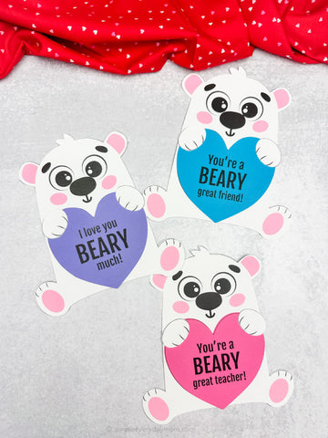 Three polar bear Valentine crafts with heart-shaped messages on a gray background with red fabric. 