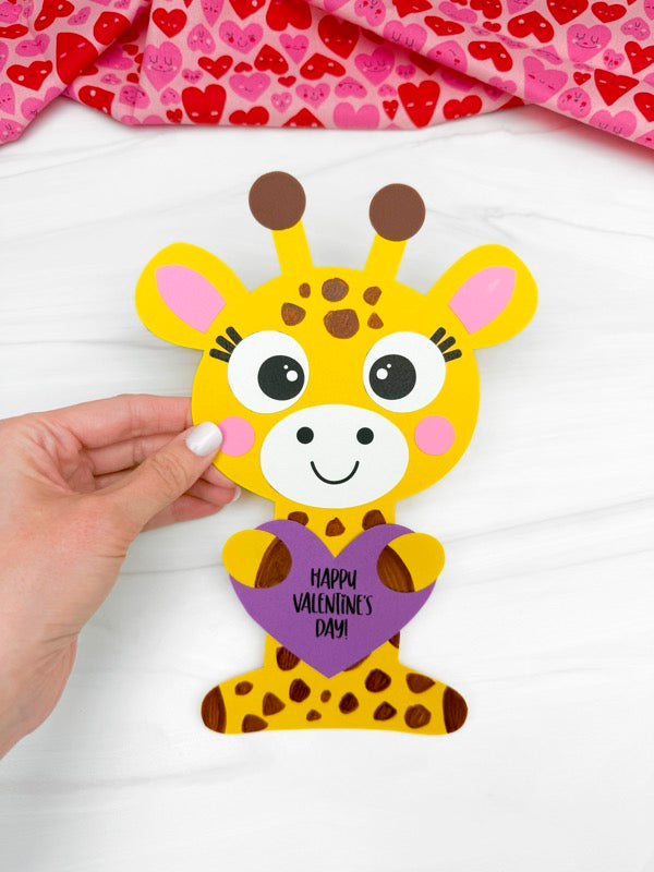 Hand holding a paper craft of a giraffe with a heart and 'Happy Valentine's Day' text on a white background.