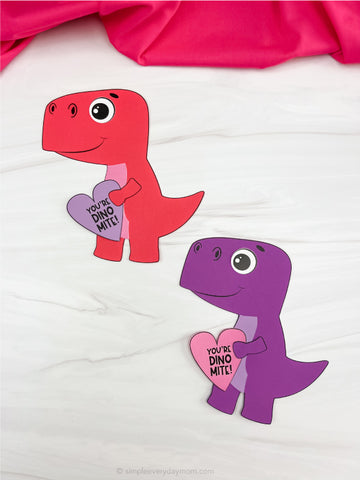 Two paper dinosaurs, one red and one purple, holding heart-shapes on a white background.