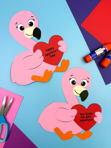 Two pink flamingo crafts holding red hearts on a colorful background with scissors and glue sticks.