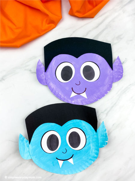 Two vampire paper plate crafts, one purple and one blue, displayed on a white marble surface with orange fabric in the background.