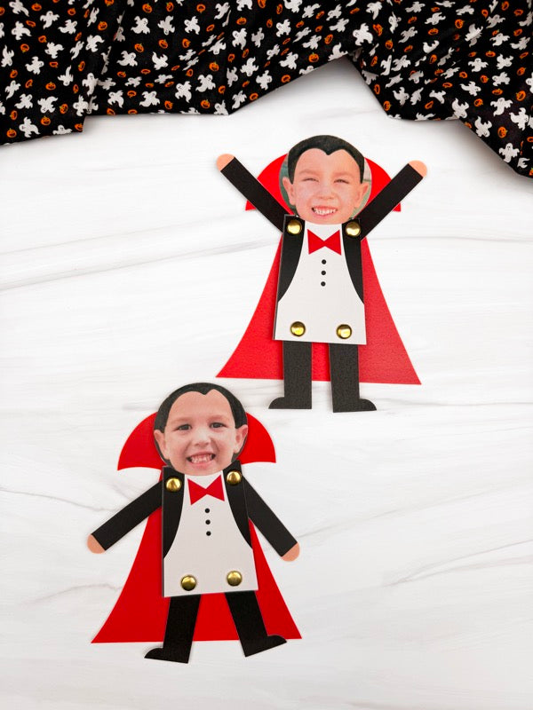 Two vampire photo crafts with printed faces, black and white tuxedo-style bodies, red cape shapes, and gold buttons, arranged on a white surface with a black-and-orange patterned fabric at the top.