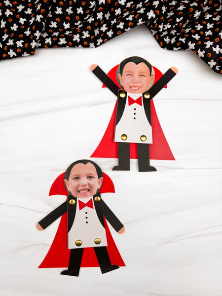 Two vampire photo crafts with printed faces, black and white tuxedo-style bodies, red cape shapes, and gold buttons, arranged on a white surface with a black-and-orange patterned fabric at the top.