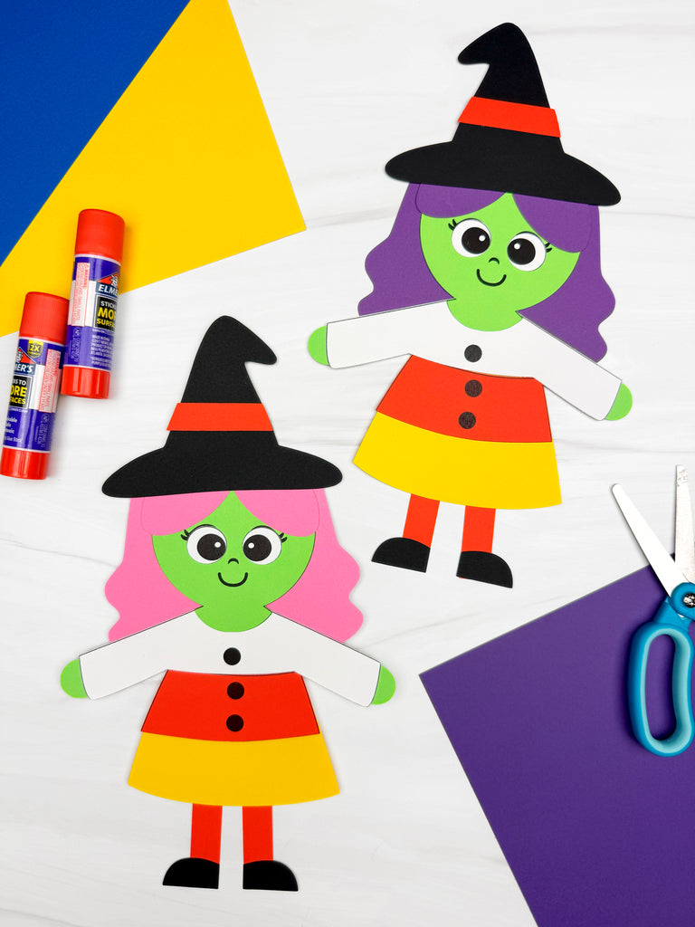 Two colorful paper witch craft figures with green faces, black hats with orange bands, and striped red, yellow, and white dresses, arranged on a white surface with craft supplies including glue sticks and scissors.