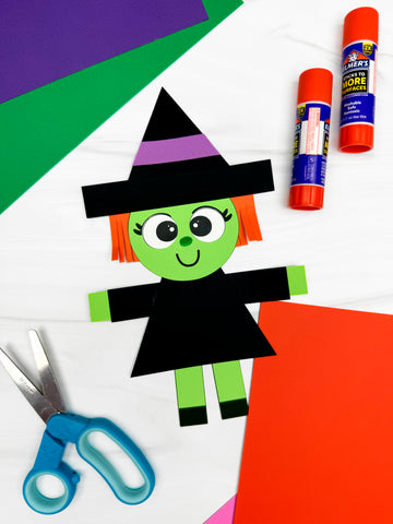 Halloween-themed paper craft with a witch, scissors, glue, and colored paper on a white background.