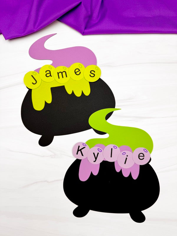 Two black cauldron-shaped name crafts, shown on a white surface with purple fabric at the top.