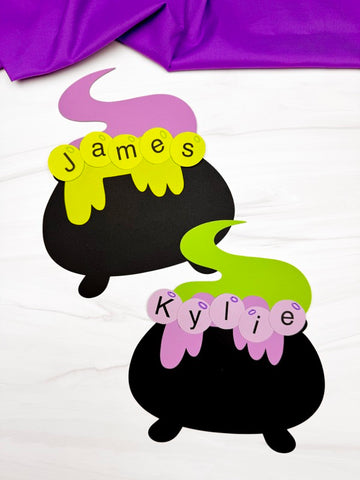 Two black cauldron-shaped name crafts, shown on a white surface with purple fabric at the top.