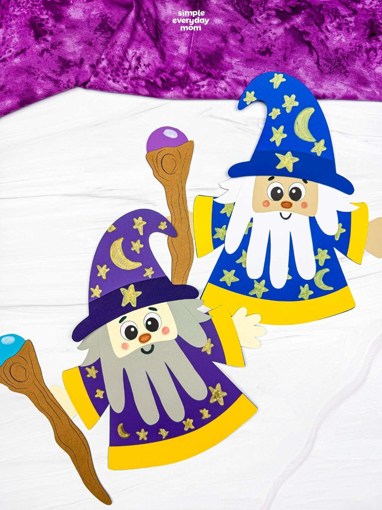 Two colorful paper wizard crafts with star-and-moon hats, long beards, and wooden staffs displayed on a white surface.