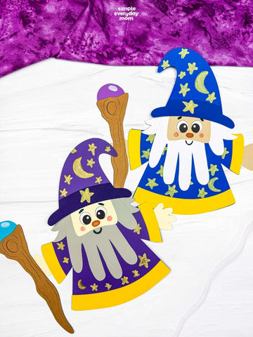 Two colorful paper wizard crafts with star-and-moon hats, long beards, and wooden staffs displayed on a white surface.