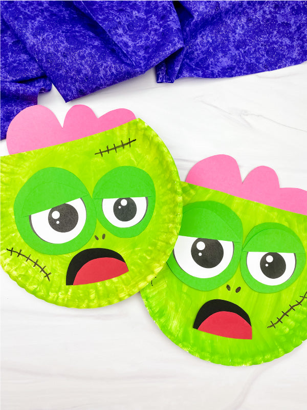 Two paper plate zombie crafts on marble background with purple fabric on top